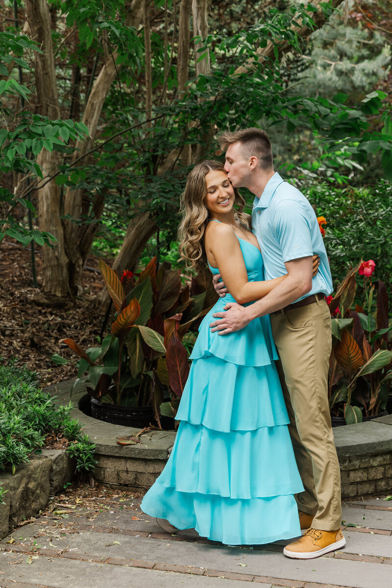 Man kissing his fiancée's forehead among tropical canna lilies at Roger Williams Botanical Gardens, she in a tiered aqua gown