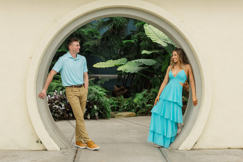 Couple smiling at each other framed in a round moon-gate archway surrounded by tropical plants at the botanical center