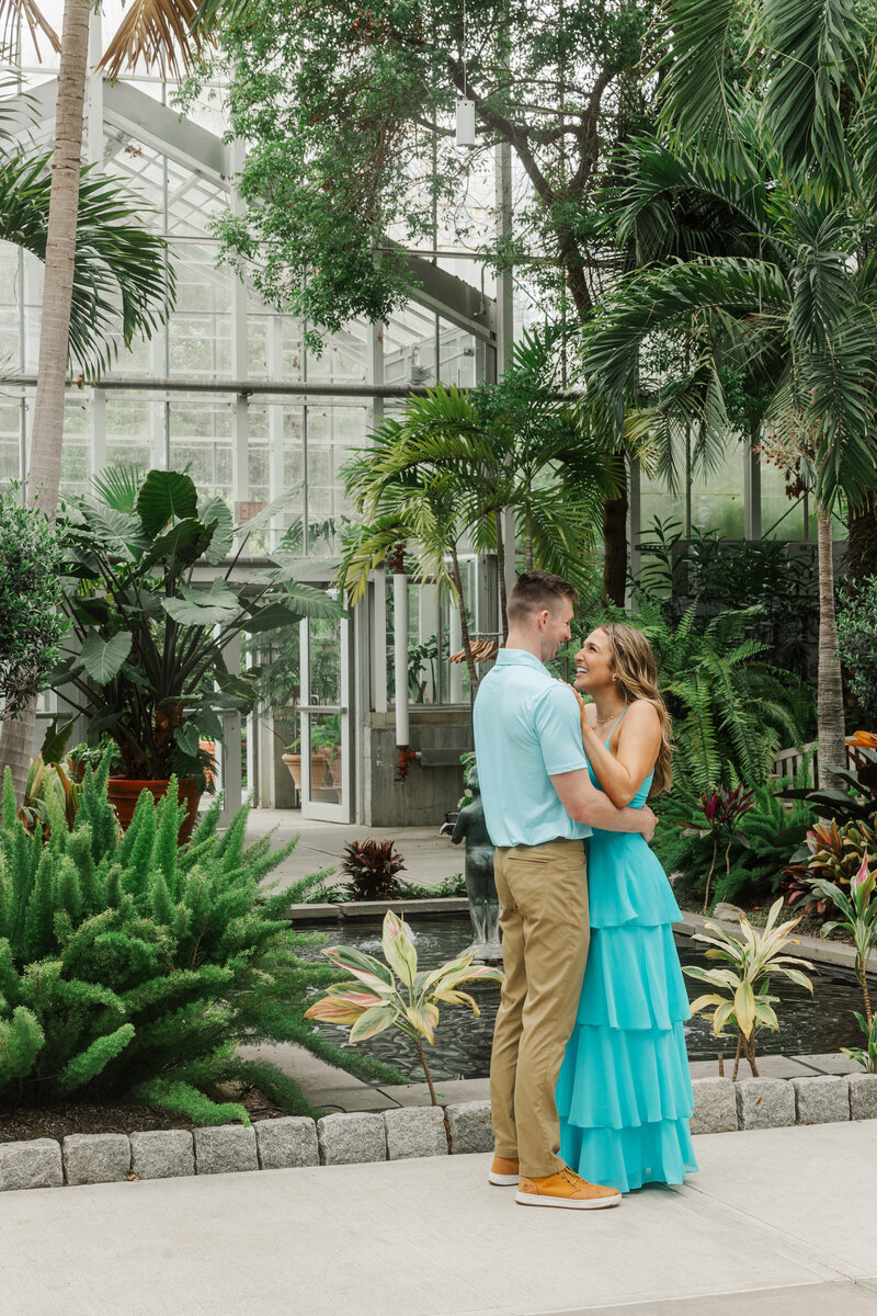 Engaged couple embracing in a glass conservatory full of palms beside a koi pond at Roger Williams Botanical Center