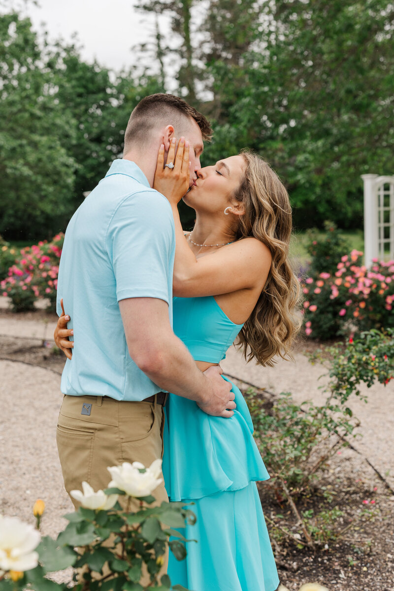 Engaged couple kissing with the woman's hand on her fiancé's cheek, colorful roses behind them