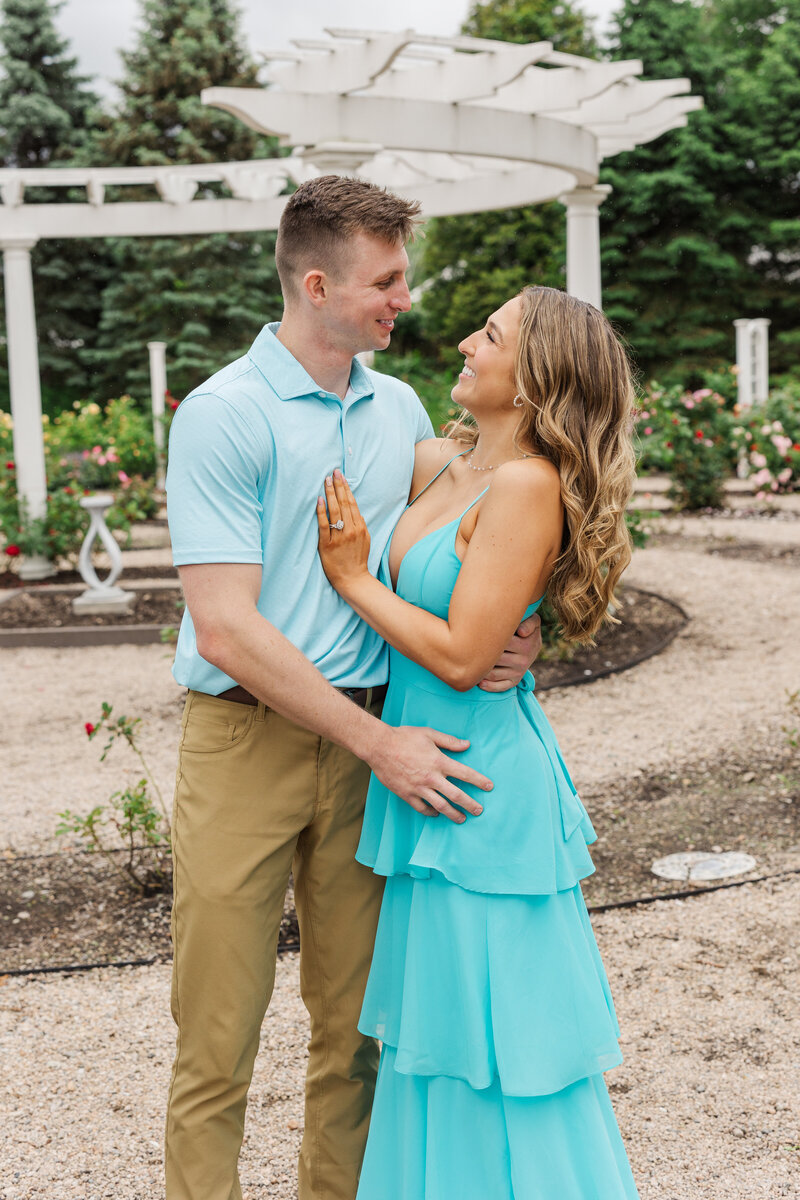 Woman resting her hand on her fiancé's chest, engagement ring showing, in a rose garden with a white pergola behind