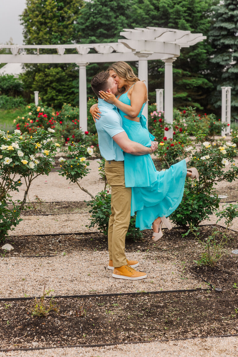 Man lifting his fiancée as they kiss among blooming roses by a white pergola at Roger Williams rose garden in Providence, RI