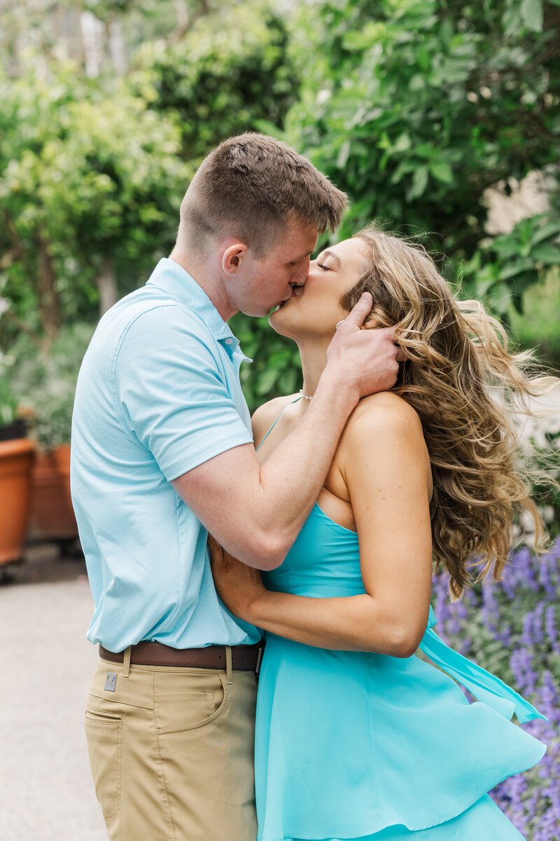 Couple kissing with the woman's hair caught in the breeze, tropical greenery and purple blooms behind