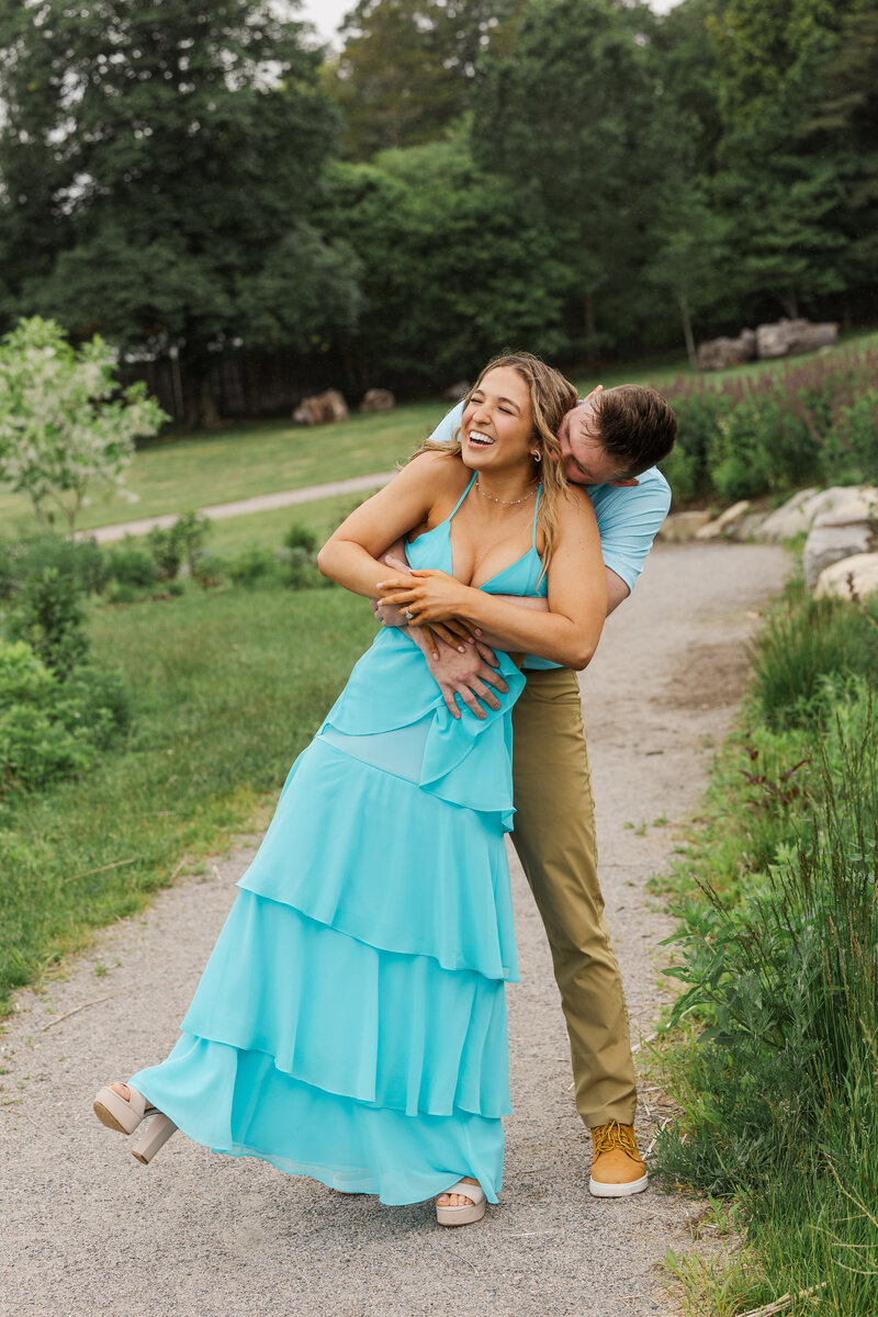 Man hugging his laughing fiancée from behind on a garden path with rocks and purple wildflowers behind