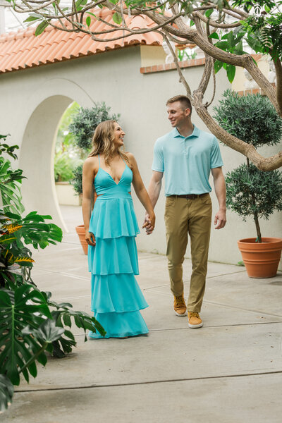Engaged couple holding hands and walking under a white moon gate past potted topiaries in a garden courtyard