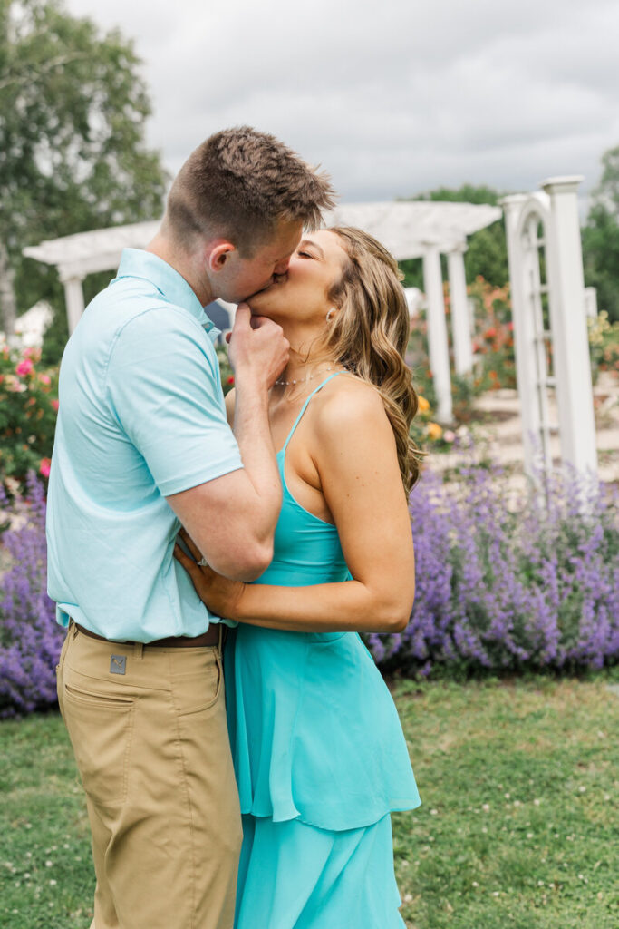 Engaged couple kissing in front of a white pergola and purple flowers in the Roger Williams rose garden