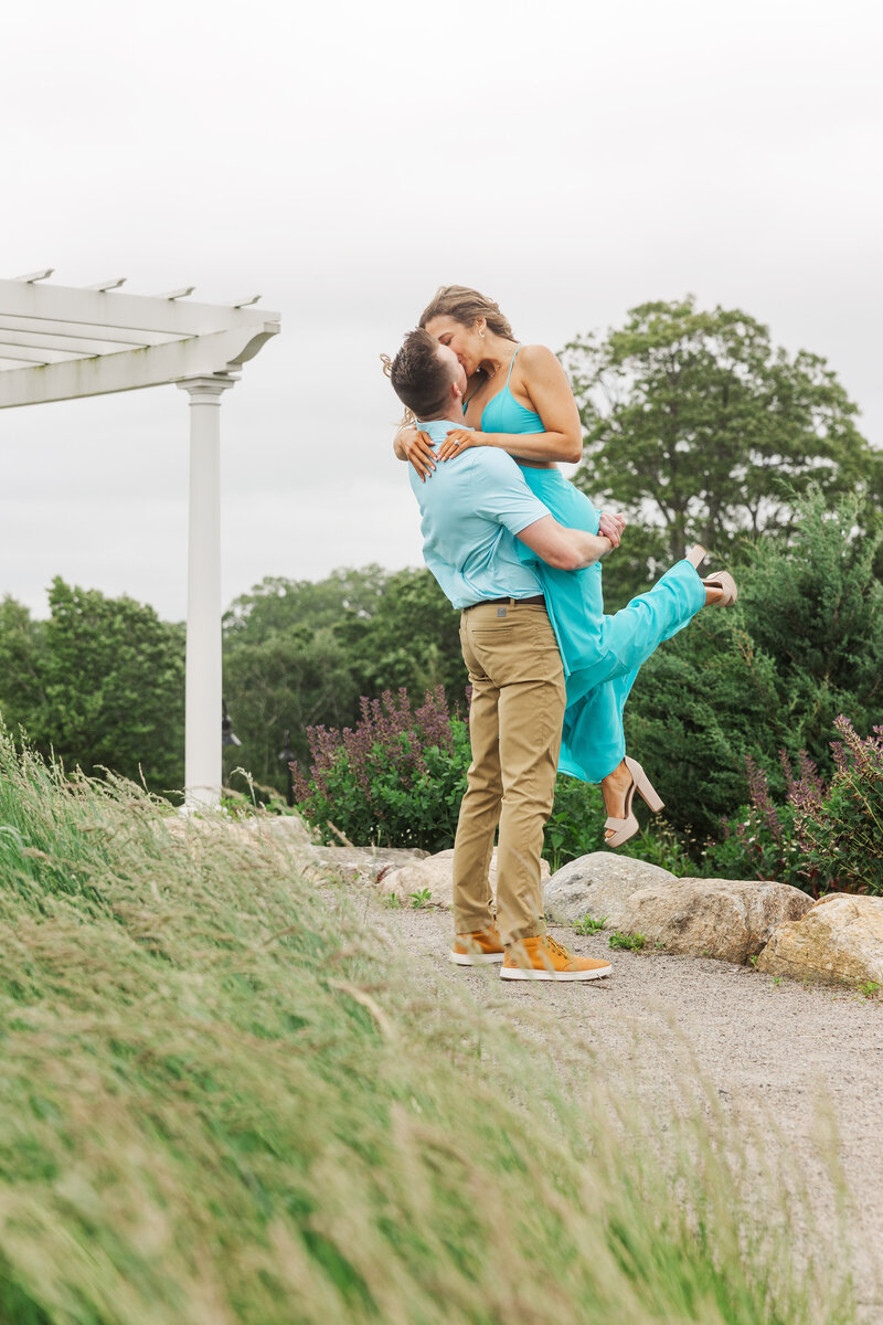Man lifting his fiancée into a kiss on a garden path beneath a white pergola under an overcast sky