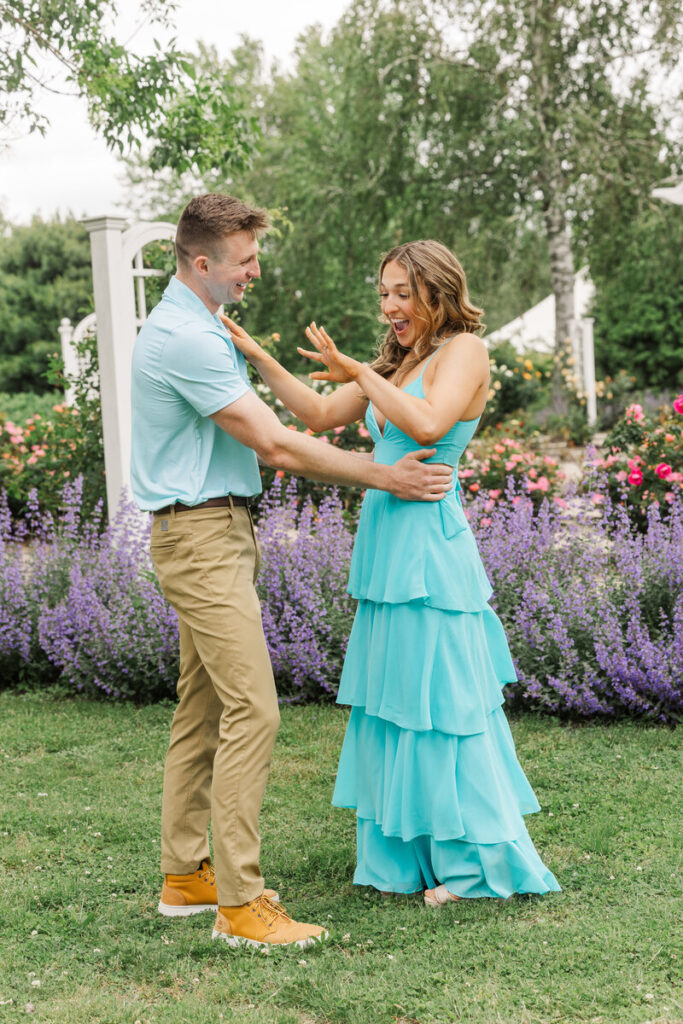 Woman reacting with hands raised in joyful surprise as her partner reaches for her in a rose and catmint garden