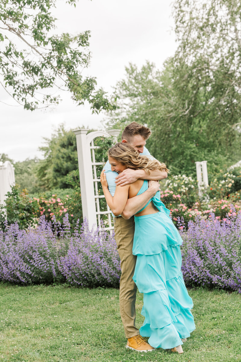 Newly engaged couple hugging tightly, the woman smiling with eyes closed, among purple catmint and roses
