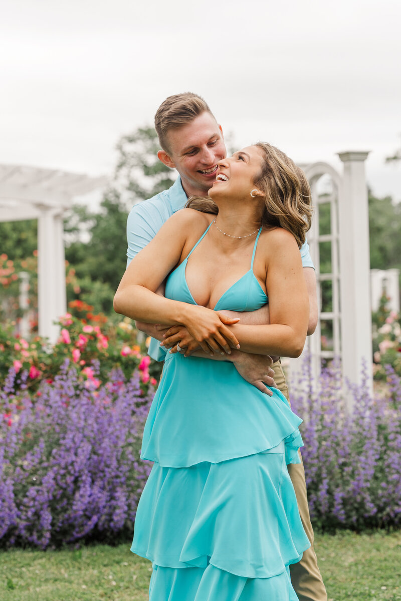 Man hugging his fiancée from behind as they both laugh in a rose garden with purple catmint and a white pergola