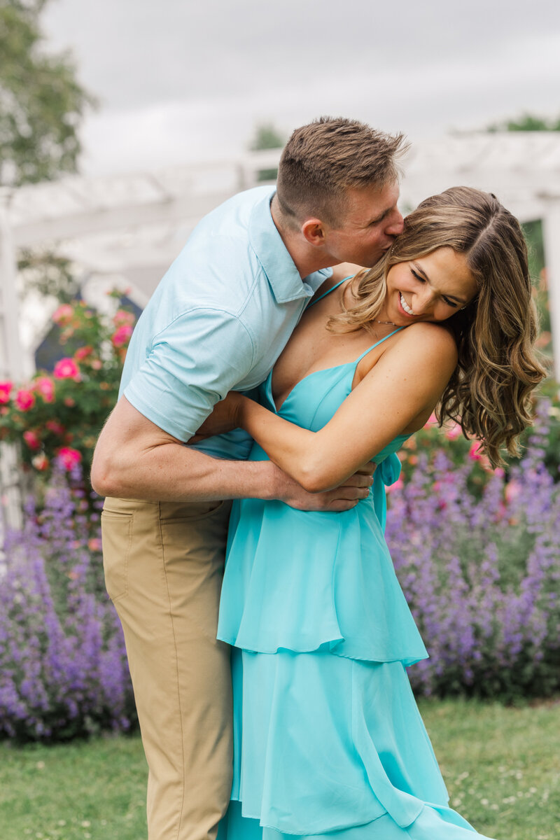 Man leaning to kiss his laughing fiancée's cheek among roses and purple catmint by a white pergola