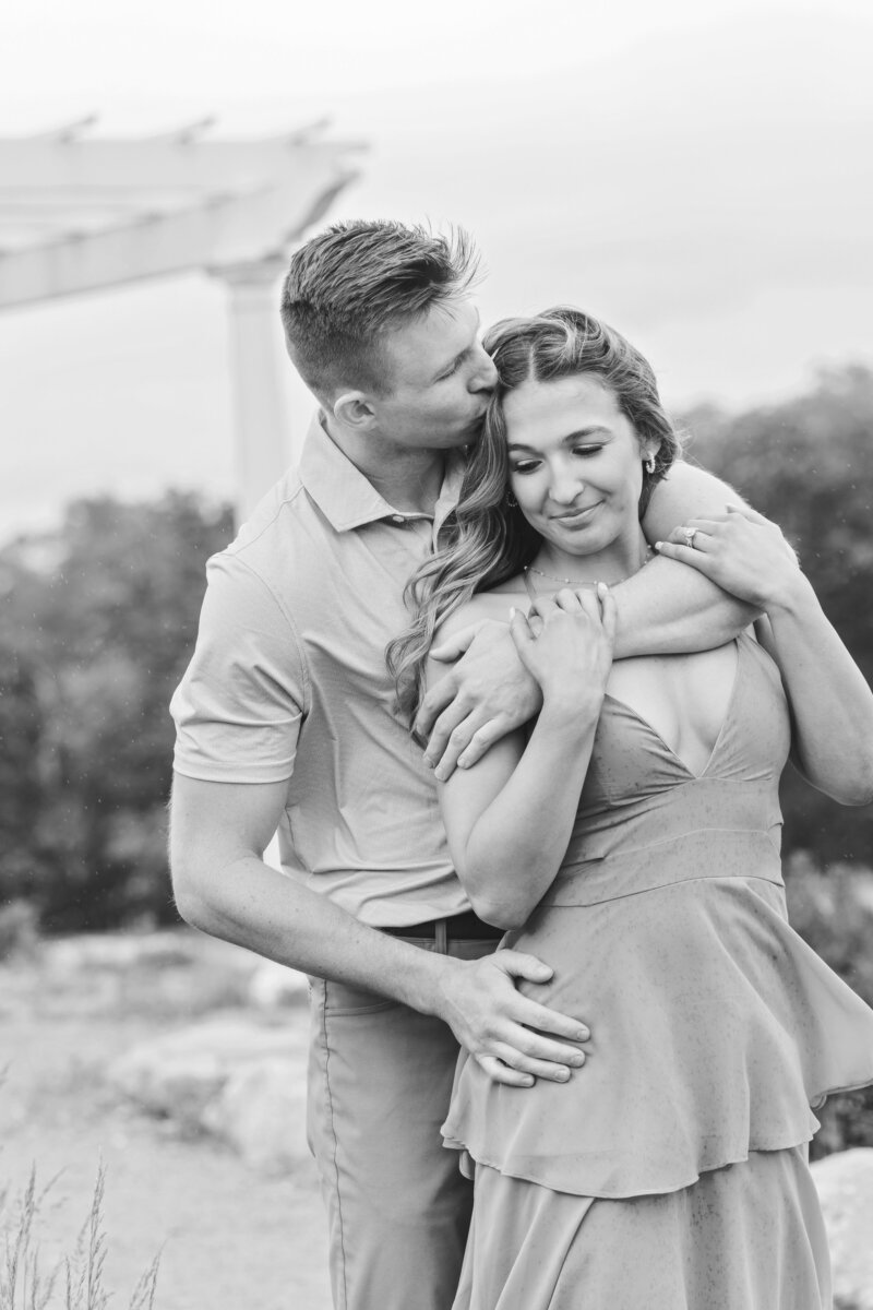 Black and white photo of a man holding his fiancée from behind and kissing her temple beneath a garden pergola