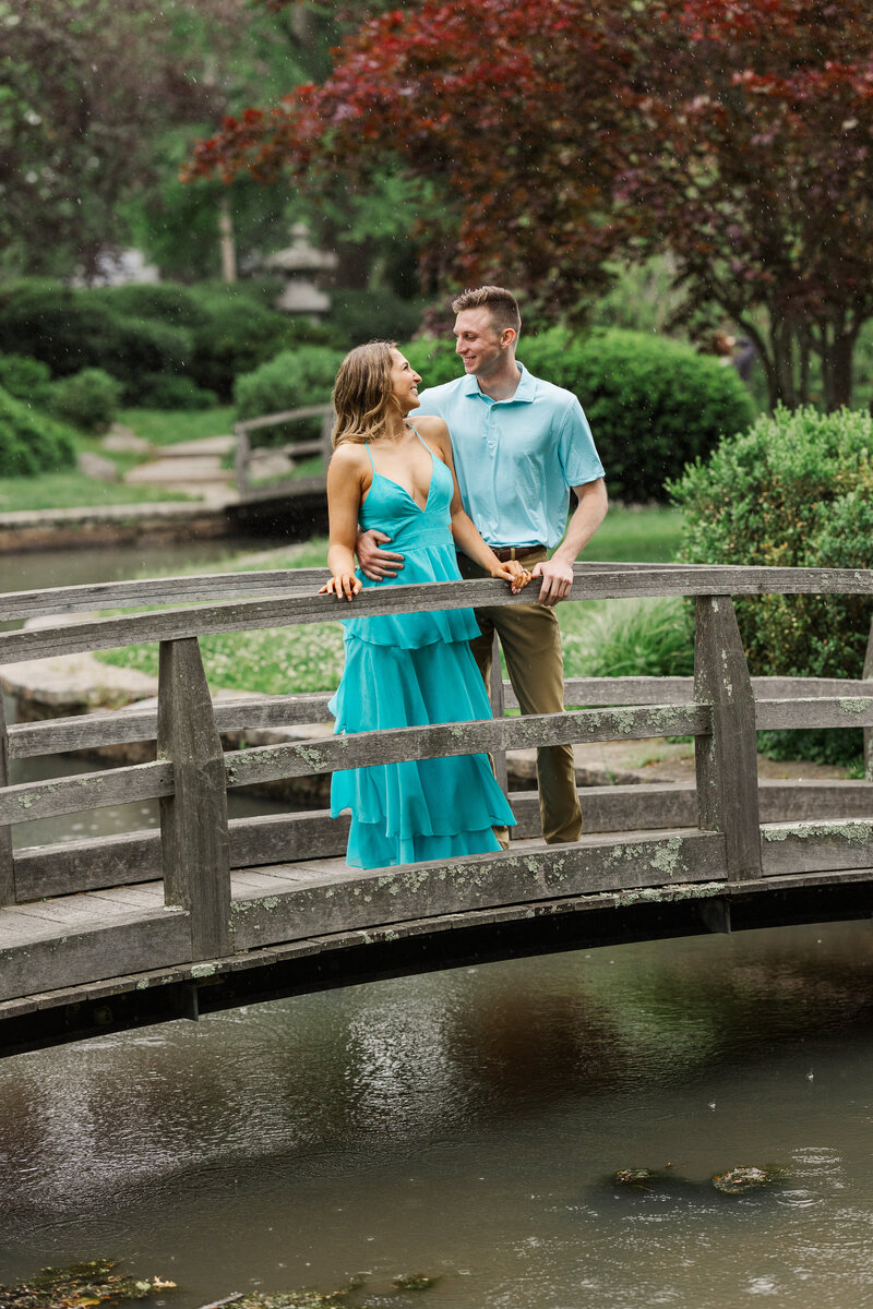 Engaged couple smiling at each other on an arched wooden bridge over a koi pond in a Japanese garden with a red maple behind