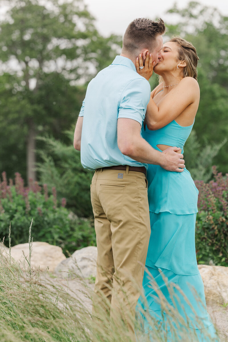 Engaged couple kissing among rocks and purple wildflowers in a light rain, the woman in an aqua cutout dress