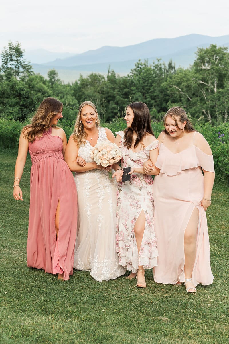 Bride laughing arm-in-arm with three bridesmaids in pink and floral dresses in a mountain meadow