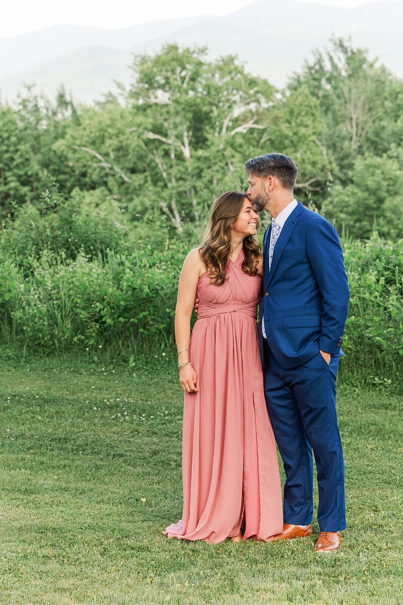 Groom in a blue suit kissing the forehead of a guest in a rose-pink dress in a grassy mountain field