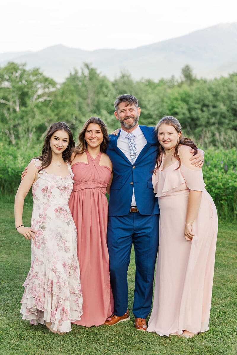 Groom in a blue suit with his three daughters in blush and floral dresses, smiling together in a green New Hampshire field