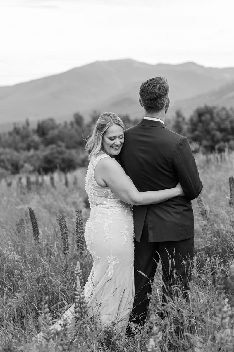 Black and white photo of a bride resting against her groom as they look out over the mountains in a tall grass field