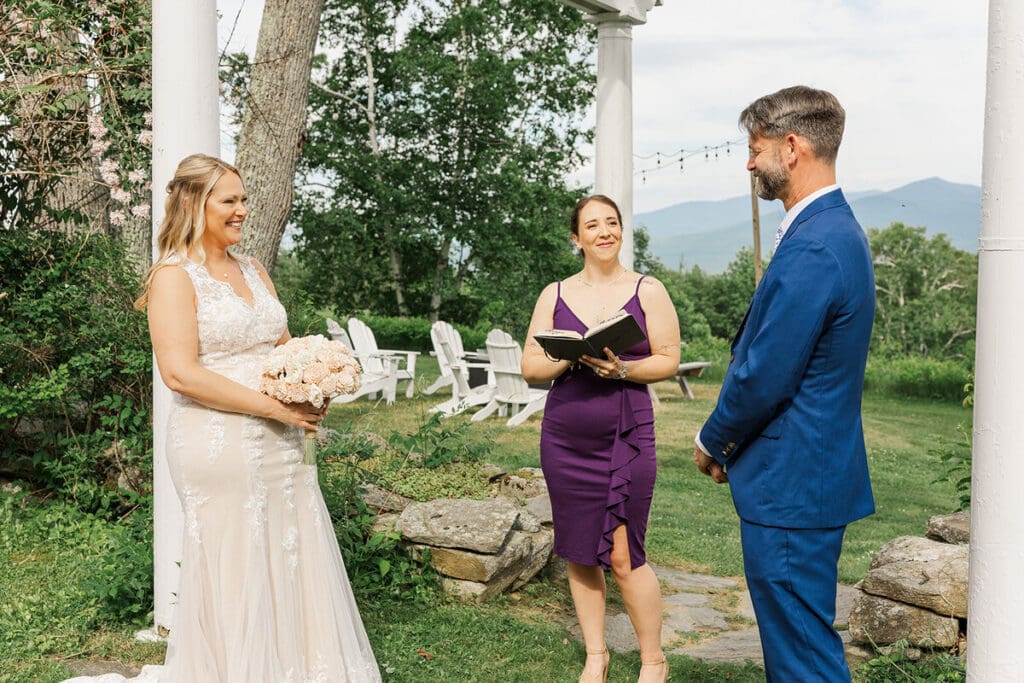 Bride and groom exchanging vows under a white arbor with a friend officiating and mountain views behind at a Sugar Hill, NH elopement
