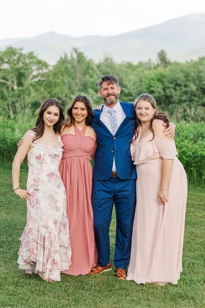 Groom in a blue suit with his three daughters in blush and floral dresses in a green field below the White Mountains