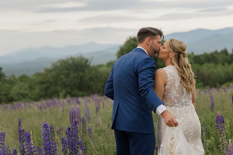 Bride and groom kissing in a Sugar Hill, NH lupine field with layered blue mountain ranges behind them