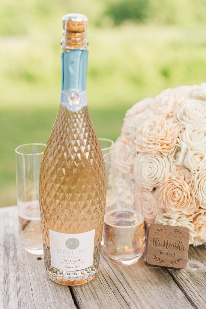Opaline brut rosé sparkling wine and two glasses beside a blush sola-wood bouquet and a box engraved "The Heaths 6-13-24"