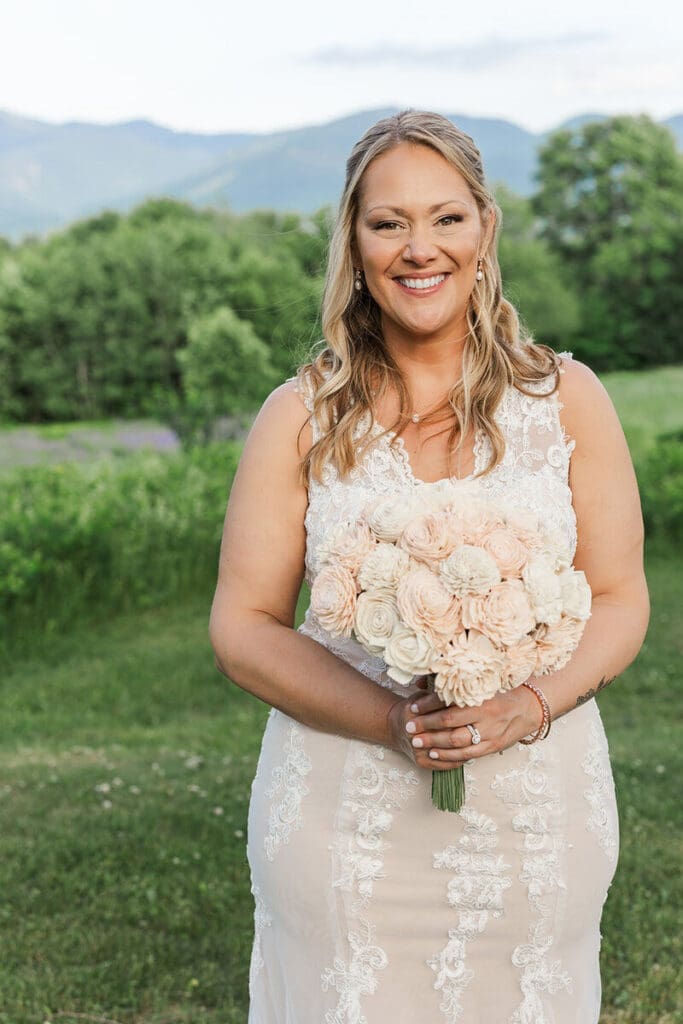 Bride in a lace gown holding a blush and ivory sola-wood bouquet, smiling with green trees and mountains behind her