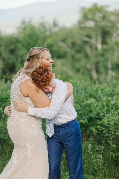 Bride hugging her young son in a white shirt and blue tie, both emotional, with green foliage behind