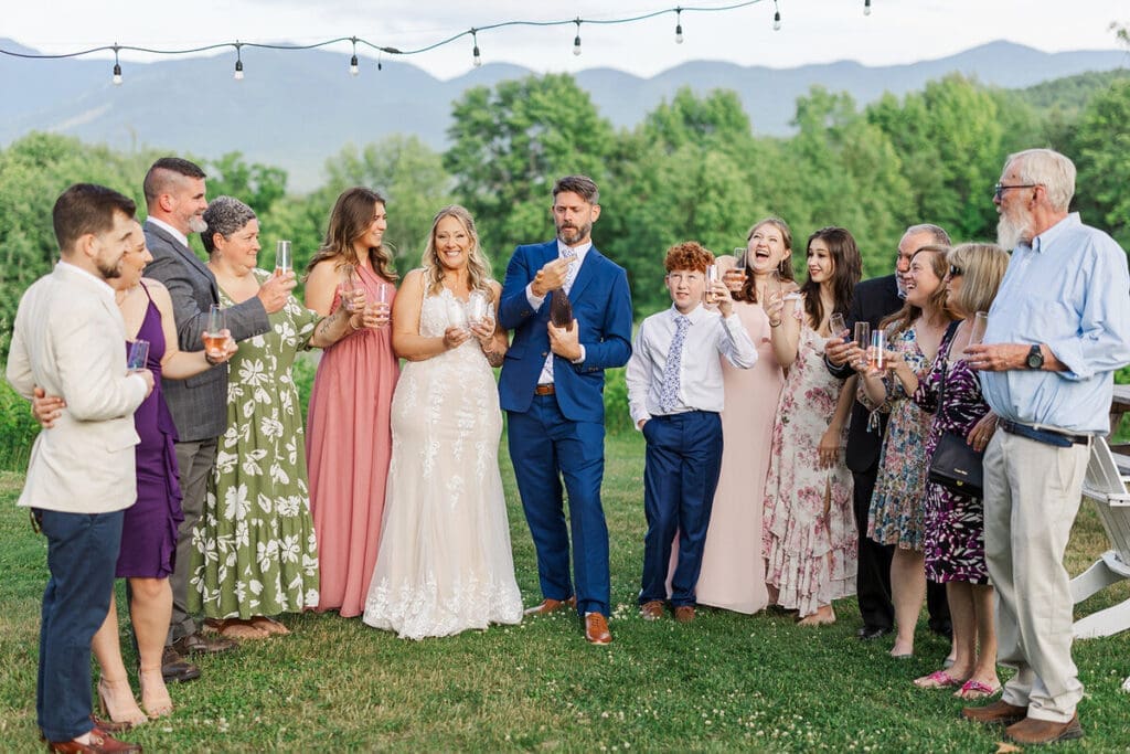 Family and guests raising a champagne toast as the groom opens a bottle, with string lights and mountain views behind
