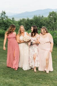 Bride laughing arm-in-arm with three bridesmaids in pink and floral dresses in a mountain meadow