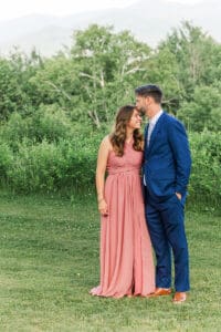 Groom in a blue suit kissing the forehead of a guest in a rose-pink dress in a grassy mountain field