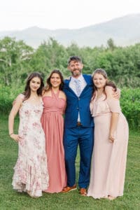 Groom in a blue suit with his three daughters in blush and floral dresses, smiling together in a green New Hampshire field
