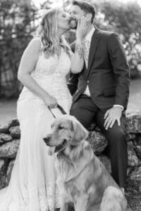Black and white photo of a bride and groom kissing on a stone wall with their golden retriever sitting in front