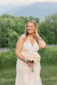 Bride with a floral arm tattoo holding a blush sola-wood bouquet, smiling near a lupine meadow