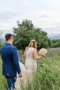 Bride and groom walking away through a lupine field toward a lone tree and mountain views, bride holding a blush bouquet