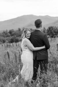Black and white photo of a bride resting against her groom as they look out over the mountains in a tall grass field