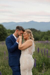 Groom kissing his bride's hand as they stand forehead to forehead in a lupine field under a soft pink sunset sky
