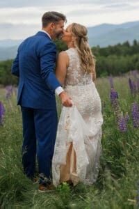 Bride and groom kissing in a lupine field, showing the lace back of her gown, with mountain views behind