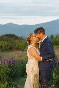 Bride and groom about to kiss in golden evening light, groom playfully holding the bouquet up, in a lupine field