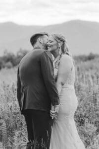 Black and white photo of a groom kissing his laughing bride's cheek in a grassy field with mountains behind