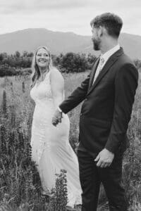 Black and white photo of a bride looking back at her groom as they hold hands walking through a Sugar Hill, NH lupine field